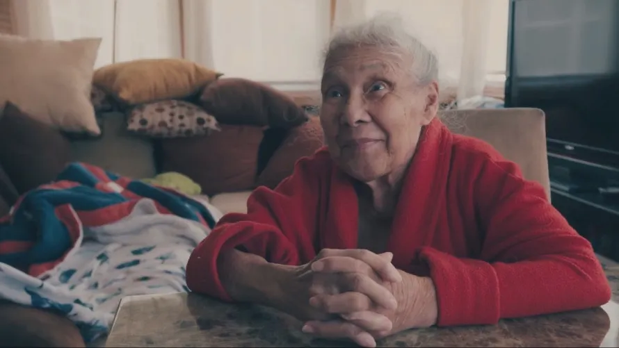 Enid & Mami festival still: an older woman in a red cardigan seated at home.